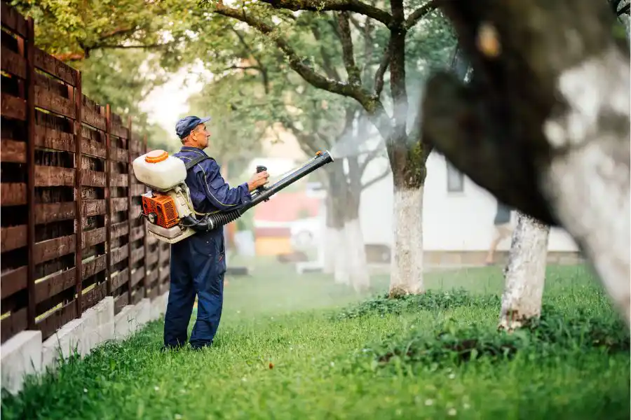 A person in a work uniform and protective gear using a backpack foliage blower to clear or maintain a grassy area near a wooden fence - keep pests away from your home with anderson pest solutions