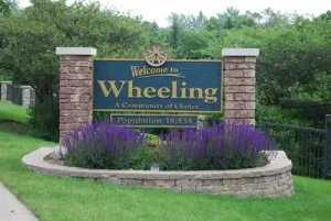 Welcome to wheeling: a community of choice – population 38,555" – a signboard at the entrance of wheeling, surrounded by greenery and vibrant purple flowers.