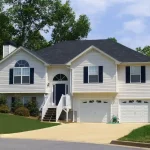 A wide shot of a classic two-story suburban house with white siding, navy blue shutters, a two-car garage, and a well-manicured green lawn under a blue sky.