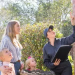 A pest control professional in a grey uniform points toward the upper corner of a brick house while a mother and two children look on.