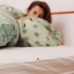 A bed bug crawling on the edge of a white mattress with a person hiding under a blanket in the background.