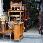 A variety of vintage and used wooden furniture, including chairs and tables, displayed on the sidewalk outside a secondhand store.