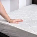 A person's hand pressing down firmly on a bare, white mattress resting on a dark bed frame.