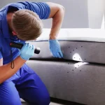 A pest control professional wearing a blue uniform and gloves using a flashlight to inspect the gap between a mattress and a grey bed base for bed bugs.