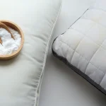 A wooden bowl of baking soda with a small wooden scoop resting next to a white quilted pillow.