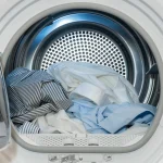 An open front-loading dryer filled with light-colored dress shirts and bedding.