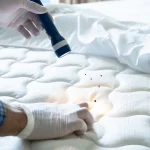 A person wearing protective gloves using a flashlight to inspect the seams of a white mattress for signs of a bed bug infestation.