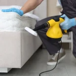 A pest control professional wearing blue gloves using a yellow handheld steam cleaner to treat the base of a mattress.