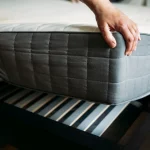 Hands lifting a mattress protected by a grey zippered encasement off a slatted wooden bed frame.