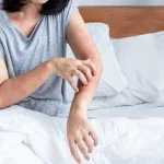 A woman sitting in bed scratching red, irritated bed bug bites on her forearm.